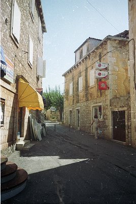 Stone-built European village street with rustic charm, featuring weathered facades and shuttered windows. A yellow awning ext...
