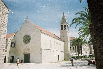 Medieval stone church with round rose window and bell tower, set in a Mediterranean courtyard. Light-colored facade contrasts...