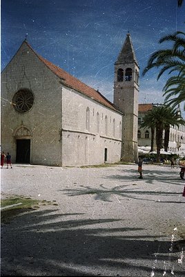 Romanesque-style church with round-arched doorway and rose window, set on a cobblestone plaza. Tall bell tower with a pointed...