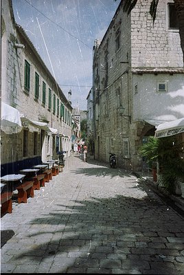 Narrow cobblestone alleyway lined with weathered stone buildings, featuring green-framed windows and outdoor seating under wh...