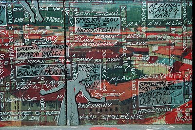 Graffiti-style mural with layered Czech/Slovak text on weathered concrete, featuring bold red and white paint. Central figure...