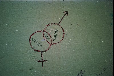 Graffiti-style initials "V" (Verca) and "M" (Martin) in red ink on a greenish surface, resembling a concrete or painted wall....