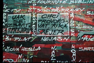 Vibrant graffiti-covered wall with faded red paint and layered stenciled text in multiple languages (Czech, Slovak, Russian)....
