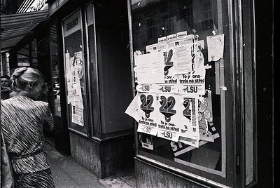 Black-and-white street scene featuring a woman in a striped dress examining posters taped to a storefront window. Prominent "...