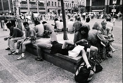 Street scene from the 1980s–1990s featuring a diverse group of people relaxing on a stone bench in a busy urban plaza. Casual...
