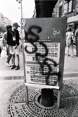 Vintage public notice board in urban setting, covered in graffiti (symbol resembling "S" or "Z"). Grid of black-and-white pho...