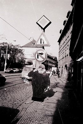 Mid-20th century urban street scene featuring a man in a long coat and hat crossing a cobblestone roadway. Traffic signs (yie...