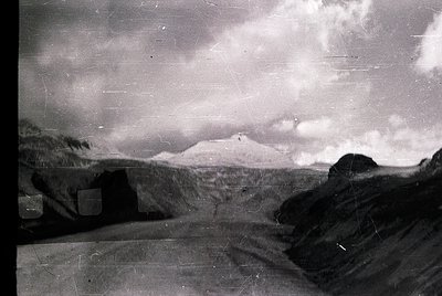 Vintage black-and-white alpine landscape with rugged terrain and distant snow-capped peak. Foreground shows weathered stone w...