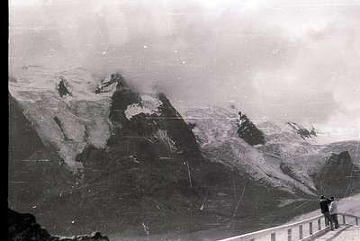 Vintage black-and-white alpine scene featuring jagged peaks and snow-covered slopes. A lone figure stands on a viewing platfo...
