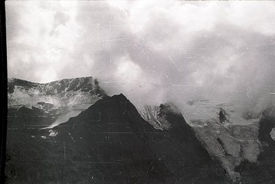 Vintage black-and-white aerial shot of rugged alpine terrain with steep peaks and mist-covered slopes. Likely mid-20th centur...