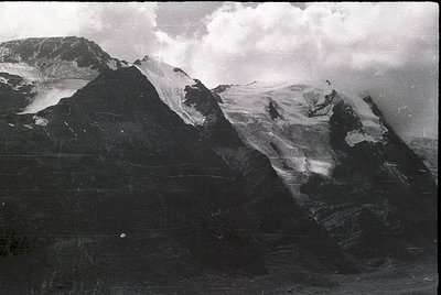 High-contrast black-and-white alpine landscape featuring jagged peaks and snowfields. Dramatic cloud cover obscures mid-mount...