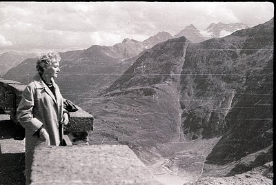 Mid-century woman in a structured coat poses atop rugged alpine ridge, framed by jagged peaks. Vintage monochrome print sugge...