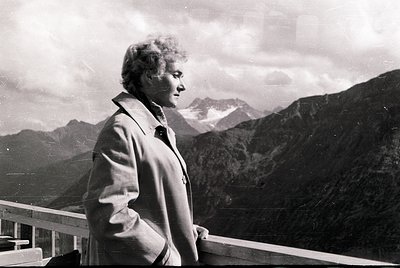 Mid-century woman in a collared coat stands on a metal railing overlooking rugged alpine peaks, suggesting mid-20th century o...