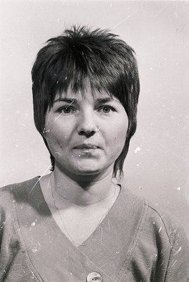 Portrait of a woman with a short, angled bob hairstyle, taken in black-and-white. She wears a simple V-neck sweater and a sma...