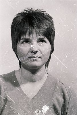 Portrait of a woman with short, straight hair and a neutral expression, captured in black-and-white. She wears a simple V-nec...