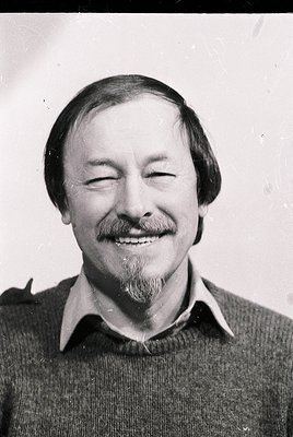 Portrait of a man with a relaxed, joyful expression—eyes closed, mustache, and short hair. Wearing a collared sweater. Black-...