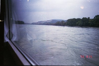 Vintage view through a train window showing turbulent river waters, likely the Danube, with distant forested hills. Overcast ...