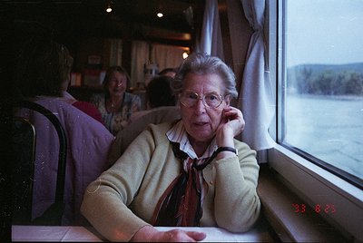 Vintage train interior shot featuring an elderly woman in a cream sweater and red scarf speaking on a corded phone, seated ne...