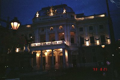 Neoclassical building illuminated at dusk, featuring arched windows, balustrades, and ornate rooftop details. Warm lighting h...