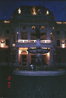 Neoclassical building façade illuminated at dusk, featuring a grand central fountain with a sculpted figure atop. Balcony wit...