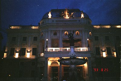 Neoclassical building illuminated at dusk, featuring ornate columns, balustrades, and a central fountain. The inscription "Sl...
