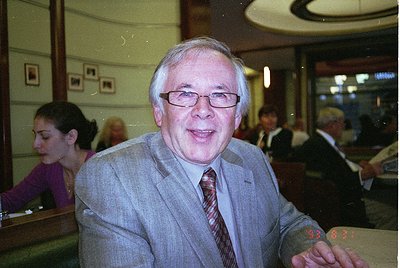Classic 1990s-era indoor portrait in a formal setting. Man in a light grey suit, red-striped tie, and glasses poses with a pl...