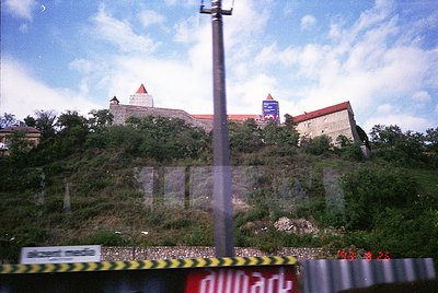 Historic hilltop fortress with medieval architecture, featuring stone walls and conical towers. Overgrown vegetation surround...