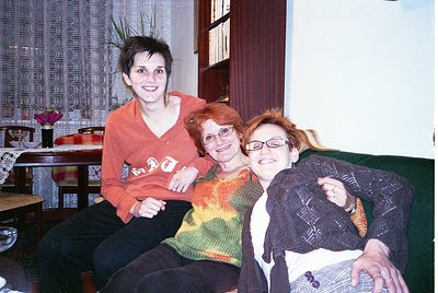 Three women pose indoors in a cozy, vintage setting—likely early 2000s. Left: woman in a red hoodie with a bold graphic. Cent...