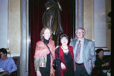 Three adults pose indoors near a grand statue in a formal setting, likely a museum or cultural venue. The woman on the left w...