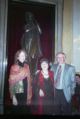Three individuals pose indoors beside a classical bronze statue of a draped figure in a grand hall, likely a museum or cultur...