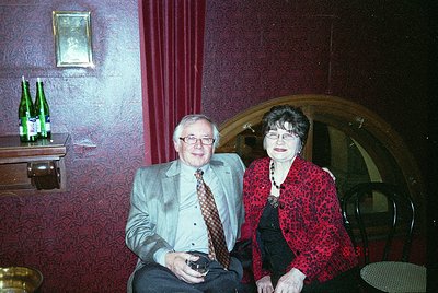 Two individuals pose indoors in a vintage setting, likely late 20th century. The man wears a light gray suit with a patterned...