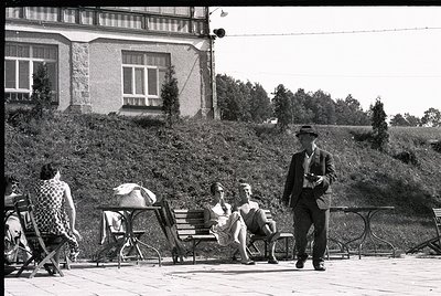 Mid-20th century urban park scene featuring vintage metal benches, stone building with large windows, and lush greenery. A ma...