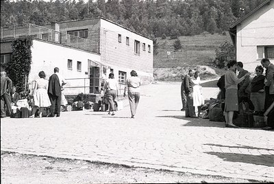 Mid-20th century rural gathering near modernist brick buildings with flat roofs and balconies. Group of people—men in suits, ...