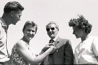 Four individuals pose outdoors in 1960s-era attire—men in suits, women in patterned dresses and hairstyles. The man in sungla...