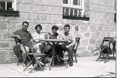 Four individuals pose outdoors at a round metal table, seated on vintage folding chairs against a stone wall with flower boxe...