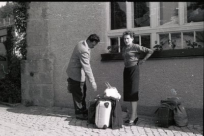 Mid-20th century street scene: man in suit and tie bends to secure a vintage suitcase, woman in structured dress leans agains...