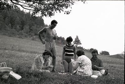 Mid-20th century family picnic in a grassy field, likely 1950s–1960s. Five individuals—two adults standing, three seated—pose...