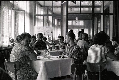Mid-century café interior with 1950s-60s women dining at round tables, clad in floral and striped dresses. Natural light floo...