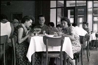 Mid-century café scene with six patrons seated at a long table, dressed in 1960s-70s attire. White tablecloths, metal chairs,...