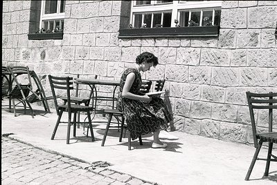 Mid-20th century woman reading outdoors at a café table, dressed in a floral dress. Stone building with flower boxes in windo...