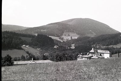 Rural alpine valley with scattered farmhouses and dense forest. Mid-20th century architecture—sloped roofs, stone foundations...