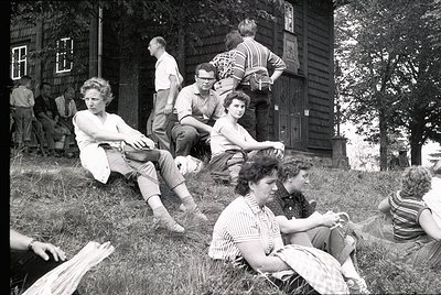 Group of young people lounging on grass near a rustic wooden house, mid-20th century. Casual summer attire—shorts, striped sh...