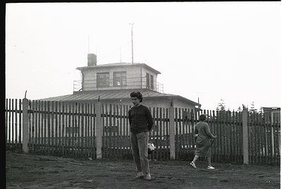 1960s-era watchtower with cylindrical observation deck, surrounded by a chain-link fence. Two individuals in casual mid-centu...