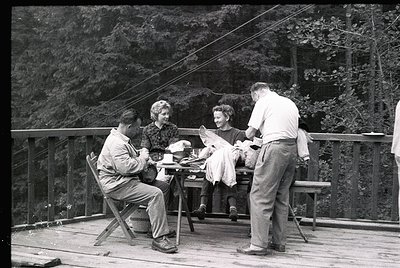 Mid-20th century group picnic on a wooden deck overlooking forested hills. Four adults seated/standing around a small table, ...