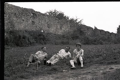 Three individuals pose casually in mid-20th-century attire—lightweight shirts, trousers, and sandals—against a weathered ston...