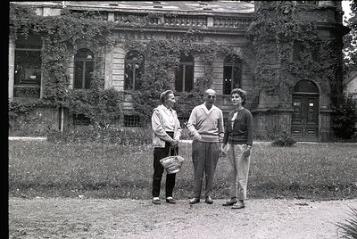 Three individuals pose outdoors in front of a grand, ivy-covered building with arched windows and ornate stonework, likely mi...