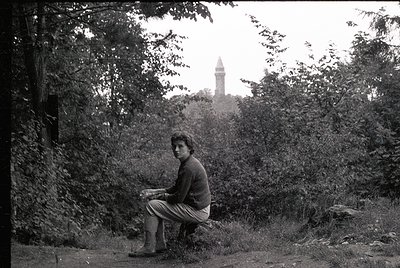 A man sits on a grassy hillside, reading a book, with dense foliage framing the shot. In the background, a tall, cylindrical ...
