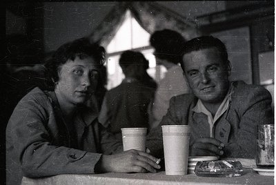 Vintage black-and-white photo of two seated individuals at a café, holding disposable paper cups. Man wears a jacket with a p...