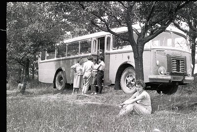 Vintage 1950s-60s scene: group of people near a parked Scania bus in a grassy, wooded area. Four adults and a seated woman in...