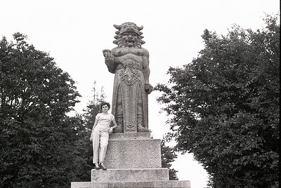 Monumental stone statue of a mythical guardian figure with bull’s head and ornate armor, holding a raised sword. Woman in 196...